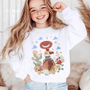 K&ouml;nnte beinhalten: Ein wei&szlig;es Langarm-Sweatshirt mit einem Grafikdruck, der eine Cowgirl mit braunem Hut auf einem Pferd zeigt, umgeben von Kakteen, bunten Blumen, blauen Wolken und Musiknoten. Das Motiv befindet sich mittig auf der Vorderseite des Kleidungsst&uuml;cks.
