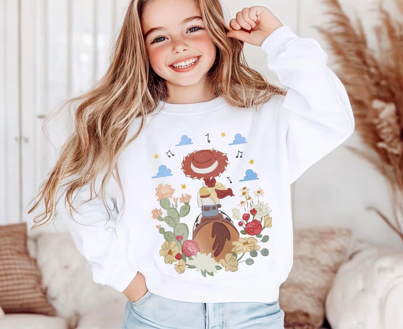 K&ouml;nnte beinhalten: Ein wei&szlig;es Langarm-Sweatshirt mit einem Grafikdruck, der eine Cowgirl mit braunem Hut auf einem Pferd zeigt, umgeben von Kakteen, bunten Blumen, blauen Wolken und Musiknoten. Das Motiv befindet sich mittig auf der Vorderseite des Kleidungsst&uuml;cks.