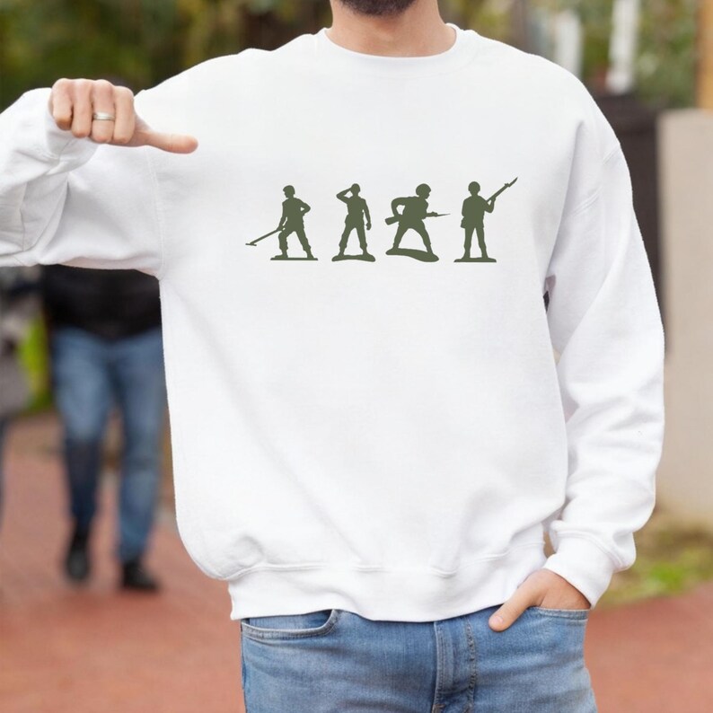May include: White sweatshirt featuring a green silhouette design of four toy soldiers in various poses. The design is centered on the front of the sweatshirt. The sweatshirt is made of a soft material.