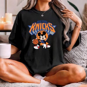 Puede incluir: Una camiseta negra de manga corta con un gr&aacute;fico de Mickey Mouse botando un bal&oacute;n de baloncesto. El dise&ntilde;o incluye el texto NEW YORK KNICKS en letras naranjas y azules sobre el personaje. La camiseta tiene cuello redondo y corte holgado.