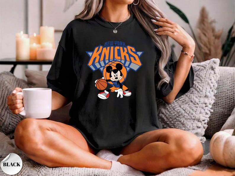 Puede incluir: Una camiseta negra de manga corta con un gr&aacute;fico de Mickey Mouse botando un bal&oacute;n de baloncesto. El dise&ntilde;o incluye el texto NEW YORK KNICKS en letras naranjas y azules sobre el personaje. La camiseta tiene cuello redondo y corte holgado.