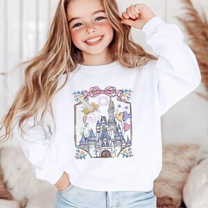 May include: White sweatshirt featuring a colorful illustration of a fairytale castle with fireworks, stars, and balloons. The design includes a pink bow and floral accents. The sweatshirt is a crew neck style.