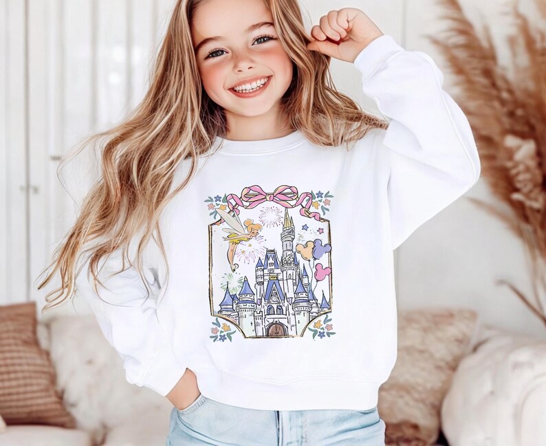 May include: White sweatshirt featuring a colorful illustration of a fairytale castle with fireworks, stars, and balloons. The design includes a pink bow and floral accents. The sweatshirt is a crew neck style.