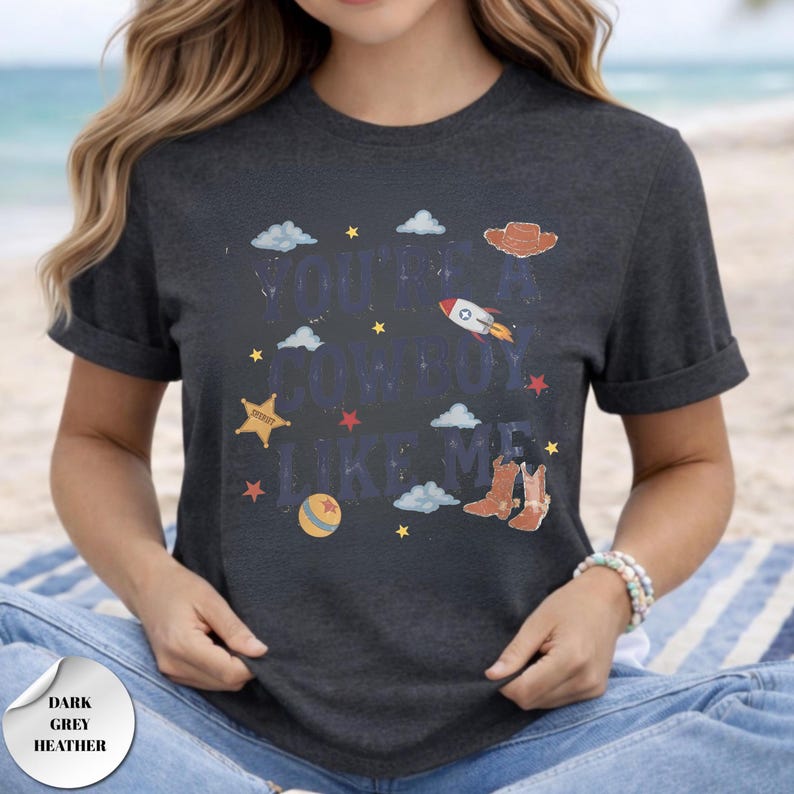 May include: A dark grey heather t-shirt featuring the text "YOU'RE A COWBOY LIKE ME" in a distressed blue font. The design includes illustrations of a brown cowboy hat, a rocket ship, a sheriff star, a ball, and cowboy boots, with small stars and clouds.