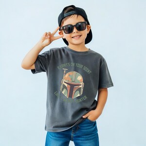 May include: A charcoal gray short-sleeve t-shirt featuring a graphic of a stylized helmet in shades of orange, brown, and cream. The text "KARMA'S ON YOUR SCENT" and "LIKE A BOUNTY HUNTER" is printed in a circular pattern around the helmet graphic.