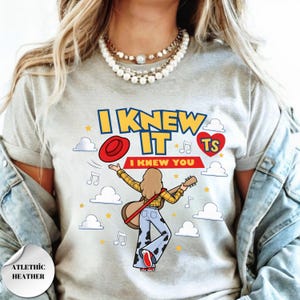 May include: A light heather gray t-shirt featuring a graphic of a person with long hair playing an acoustic guitar, wearing a yellow plaid shirt and cow-print bell-bottom pants. The text reads I KNEW IT I KNEW YOU TS with clouds, stars, and music notes.