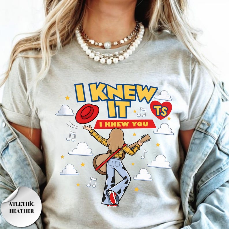 May include: A light heather gray t-shirt featuring a graphic of a person with long hair playing an acoustic guitar, wearing a yellow plaid shirt and cow-print bell-bottom pants. The text reads I KNEW IT I KNEW YOU TS with clouds, stars, and music notes.