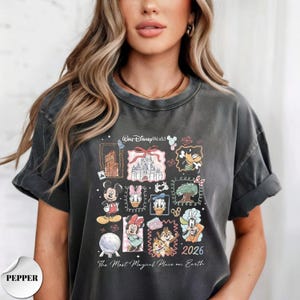 K&ouml;nnte beinhalten: Dunkelgraues T-Shirt mit einer farbenfrohen Grafik von Disney-Figuren und Wahrzeichen. Das Design zeigt Micky Maus, Donald Duck und Aschenputtels Schloss mit dem Text "Walt Disney World" und "2026". Das Shirt ist ein bequemer, l&auml;ssiger Stil.