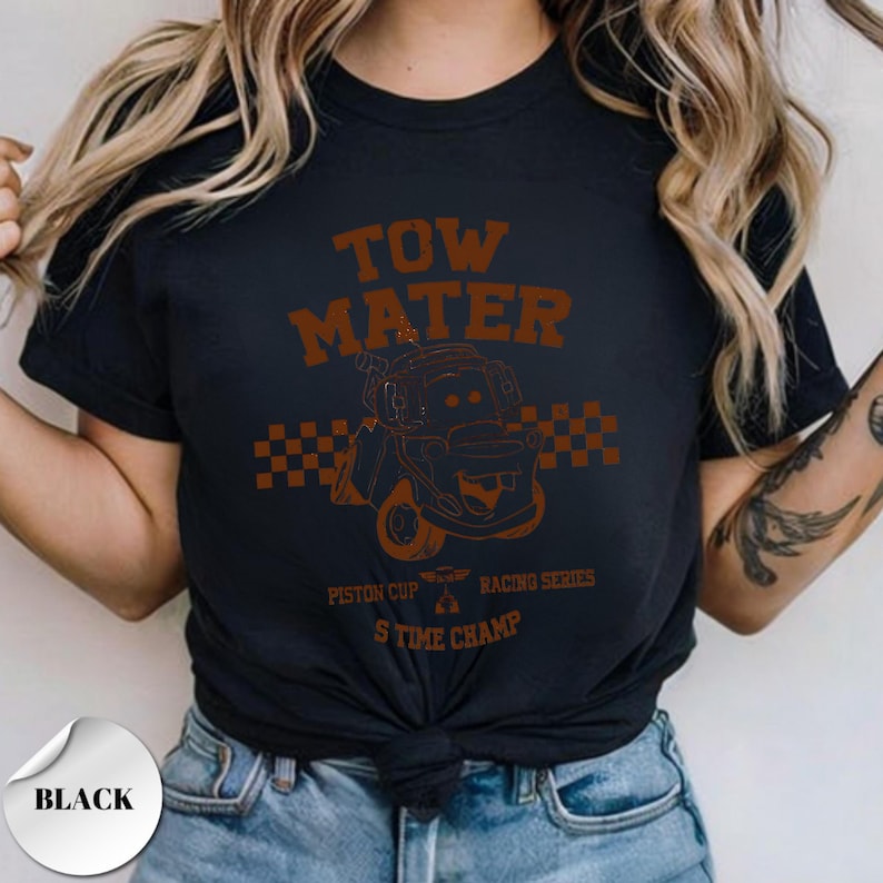 May include: A black short-sleeve t-shirt featuring a brown graphic of the character Tow Mater. The design includes a checkered flag pattern, the text TOW MATER, PISTON CUP RACING SERIES, and 5 TIME CHAMP, all printed in a distressed brown font.