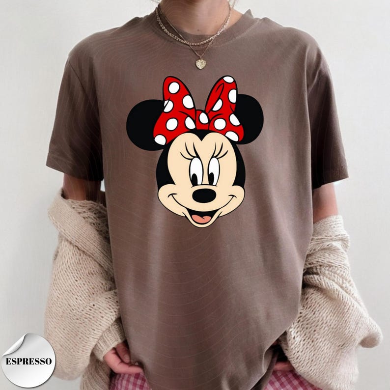 Op de afbeelding: Espresso-kleurig T-shirt met een afbeelding van Minnie Mouse's gezicht. Minnie's gezicht is zwart en bruin met een rode strik met witte stippen. Het shirt is een casual stijl.