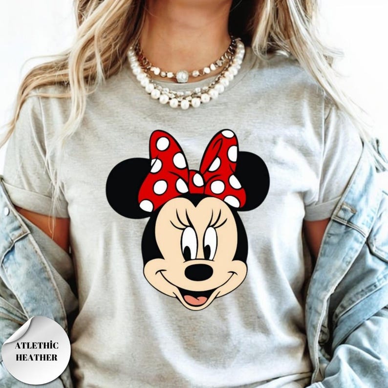 Puede incluir: Camiseta gris claro con un gr&aacute;fico de Minnie Mouse. Minnie Mouse tiene un lazo rojo con lunares blancos. La camiseta tiene las palabras "ATLETHIC HEATHER" impresas en un c&iacute;rculo blanco.