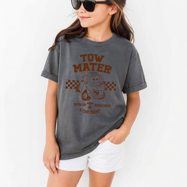 May include: A charcoal gray short-sleeve t-shirt featuring a brown graphic of the character Tow Mater from the movie Cars. The design includes the text TOW MATER, PISTON CUP RACING SERIES, and TIME CHAMP, along with a checkered pattern background.