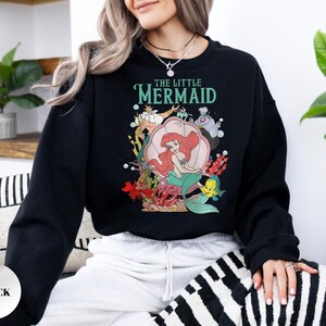 May include: A black long-sleeve sweatshirt featuring a graphic print of The Little Mermaid characters including Ariel, King Triton, Ursula, and Flounder. The text THE LITTLE MERMAID is printed in teal above the characters, with a large pink seashell.