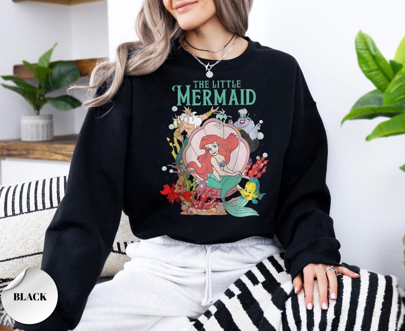 May include: A black long-sleeve sweatshirt featuring a graphic print of The Little Mermaid characters including Ariel, King Triton, Ursula, and Flounder. The text THE LITTLE MERMAID is printed in teal above the characters, with a large pink seashell.