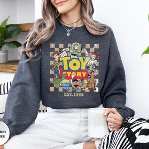 May include: A dark grey heather crewneck sweatshirt featuring a graphic print of Toy Story characters including Woody, Buzz Lightyear, Jessie, Rex, Slinky Dog, Mr. Potato Head, and an alien. The design includes a checkered background and the text Toy Story EST. 1995.