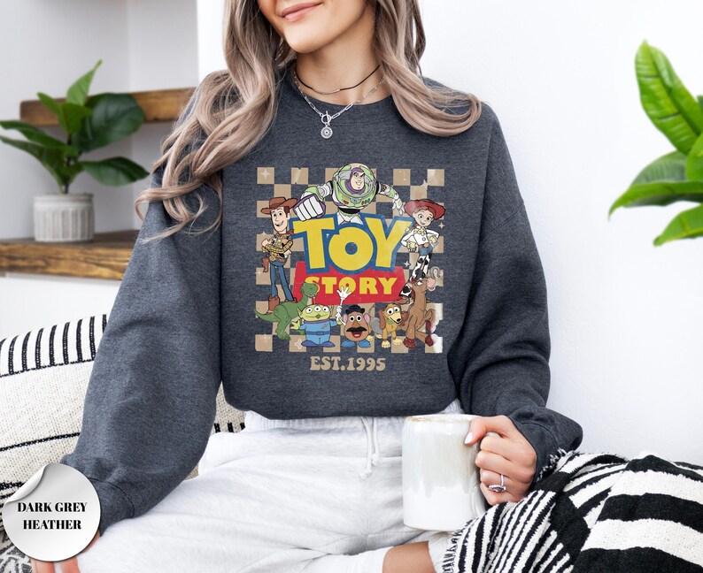 May include: A dark grey heather crewneck sweatshirt featuring a graphic print of Toy Story characters including Woody, Buzz Lightyear, Jessie, Rex, Slinky Dog, Mr. Potato Head, and an alien. The design includes a checkered background and the text Toy Story EST. 1995.