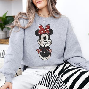 May include: A light grey sweatshirt featuring a vintage-style Minnie Mouse graphic. Minnie is depicted in black, red, and white, with a red polka-dot bow. The word "GREY" is printed on a grey circle in the lower left corner.