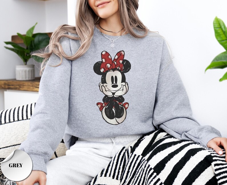 May include: A light grey sweatshirt featuring a vintage-style Minnie Mouse graphic. Minnie is depicted in black, red, and white, with a red polka-dot bow. The word "GREY" is printed on a grey circle in the lower left corner.