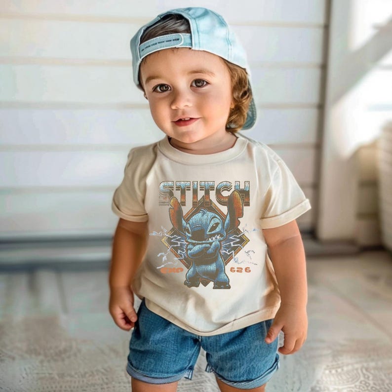 May include: A light beige short-sleeved t-shirt featuring a graphic of the blue character Stitch with the text STITCH and 626, paired with a light blue baseball cap and blue denim shorts. The graphic includes lightning bolt accents.