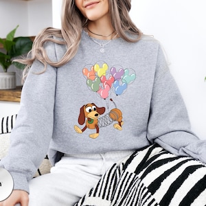 May include: A light grey sweatshirt featuring a cartoon dachshund dog character with a slinky body, holding a bunch of Mickey Mouse-shaped balloons in various colors. The word "GREY" is printed on a grey circle at the bottom.