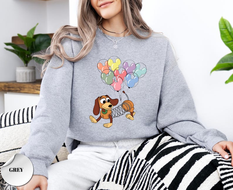 May include: A light grey sweatshirt featuring a cartoon dachshund dog character with a slinky body, holding a bunch of Mickey Mouse-shaped balloons in various colors. The word "GREY" is printed on a grey circle at the bottom.