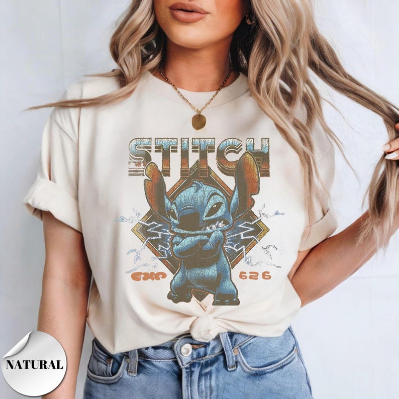 May include: A cream-colored short-sleeve t-shirt featuring a graphic of the blue character Stitch with arms crossed. The design includes the text STITCH at the top, EXP 626 at the bottom, and a diamond-shaped background with lightning bolt accents.