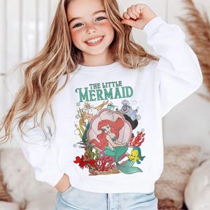 May include: A white long-sleeve sweatshirt featuring a colorful graphic print of The Little Mermaid characters, including Ariel sitting in a shell, King Triton, Ursula, and Flounder, with the text The Little Mermaid printed in green above the scene.