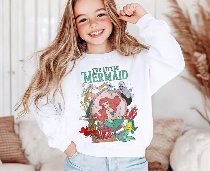 May include: A white long-sleeve sweatshirt featuring a colorful graphic print of The Little Mermaid characters, including Ariel sitting in a shell, King Triton, Ursula, and Flounder, with the text The Little Mermaid printed in green above the scene.