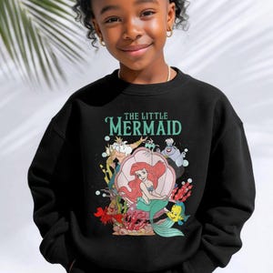 May include: A black long-sleeve sweatshirt featuring a graphic print of The Little Mermaid characters including Ariel, King Triton, Ursula, Sebastian, and Flounder. The text The Little Mermaid is printed in teal above the colorful underwater scene.