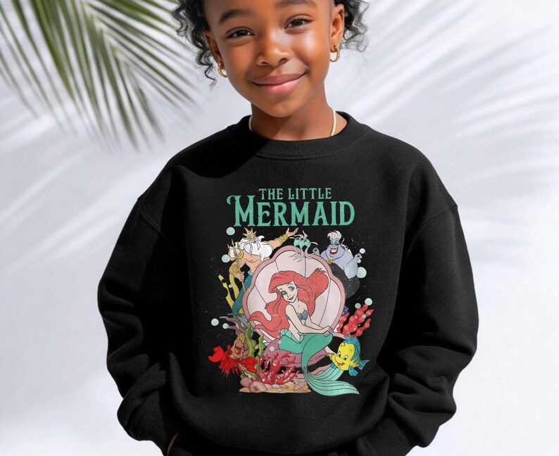 May include: A black long-sleeve sweatshirt featuring a graphic print of The Little Mermaid characters including Ariel, King Triton, Ursula, Sebastian, and Flounder. The text The Little Mermaid is printed in teal above the colorful underwater scene.