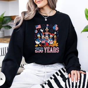 May include: A black long-sleeve crewneck sweatshirt featuring a graphic print of cartoon characters dressed in patriotic red, white, and blue clothing. The design includes the text 250 YEARS in a flag-patterned font, surrounded by balloons and fireworks.