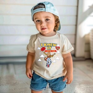 May include: A beige short-sleeved t-shirt featuring a graphic print with the text I KNEW IT and I KNEW YOU, a red hat, a red heart with TS, musical notes, clouds, and an illustration of a person playing a guitar while wearing patterned pants.