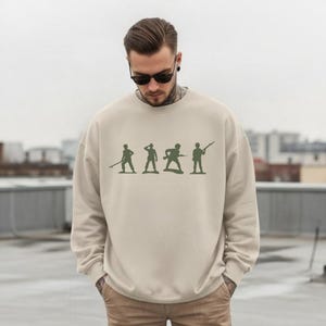 May include: A beige sweatshirt featuring a green silhouette design of toy soldiers in various poses. The long-sleeved crewneck top is made of a soft material and is suitable for casual wear.