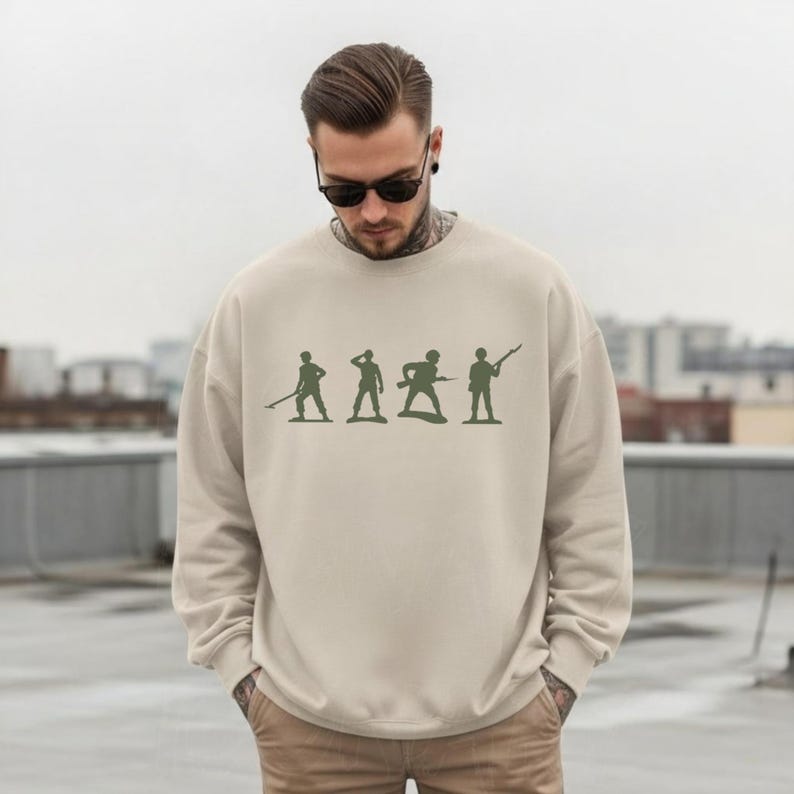 May include: A beige sweatshirt featuring a green silhouette design of toy soldiers in various poses. The long-sleeved crewneck top is made of a soft material and is suitable for casual wear.