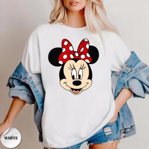 Op de afbeelding: Wit T-shirt met een grote afbeelding van Minnie Mouse's gezicht. Minnie Mouse heeft zwarte oren, een rode strik met witte stippen en een glimlachende uitdrukking. Het woord "WHITE" staat op een witte cirkel linksonder.
