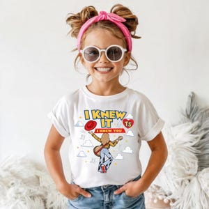 May include: A white short-sleeve t-shirt featuring a graphic of a person playing a guitar, surrounded by clouds, stars, and musical notes. The text reads I KNEW IT and I KNEW YOU, with a red hat and a red heart containing the letters TS.