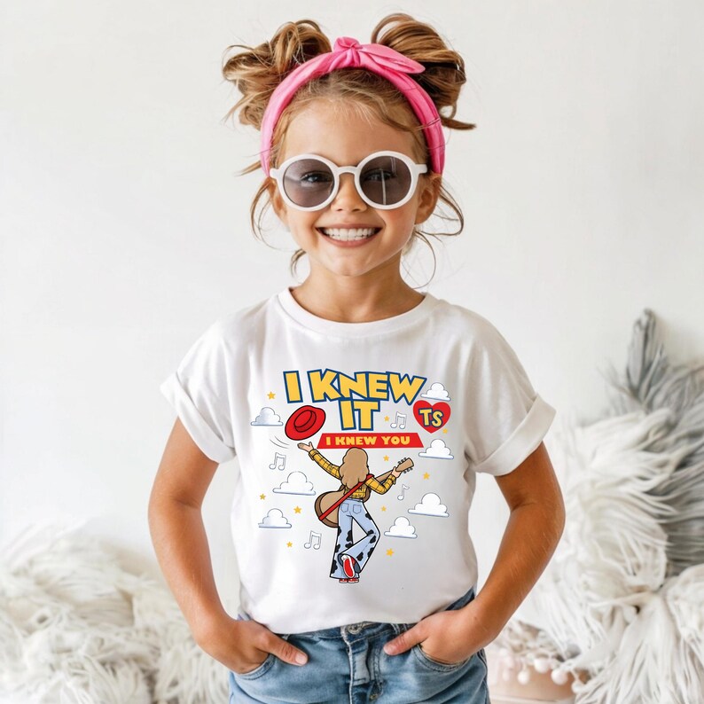 May include: A white short-sleeve t-shirt featuring a graphic of a person playing a guitar, surrounded by clouds, stars, and musical notes. The text reads I KNEW IT and I KNEW YOU, with a red hat and a red heart containing the letters TS.