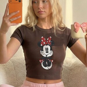May include: A brown crop top featuring a Minnie Mouse graphic with a red and white polka dot bow. The cartoon character is in a seated pose. The top has short sleeves.