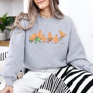 Op de afbeelding: Een lichtgrijs sweatshirt met een cartoonontwerp van figuren die oranje verkeerskegels dragen. De figuren staan in een rij, met de kegels op hun hoofd. Het sweatshirt is effen van kleur.