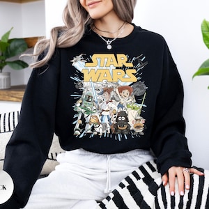 Puede incluir: Una sudadera negra de manga larga con un estampado gr&aacute;fico de personajes de Star Wars reimaginados como figuras de Toy Story. El dise&ntilde;o incluye el logotipo amarillo de Star Wars sobre ilustraciones de personajes como Woody, Jessie y Darth Vader.