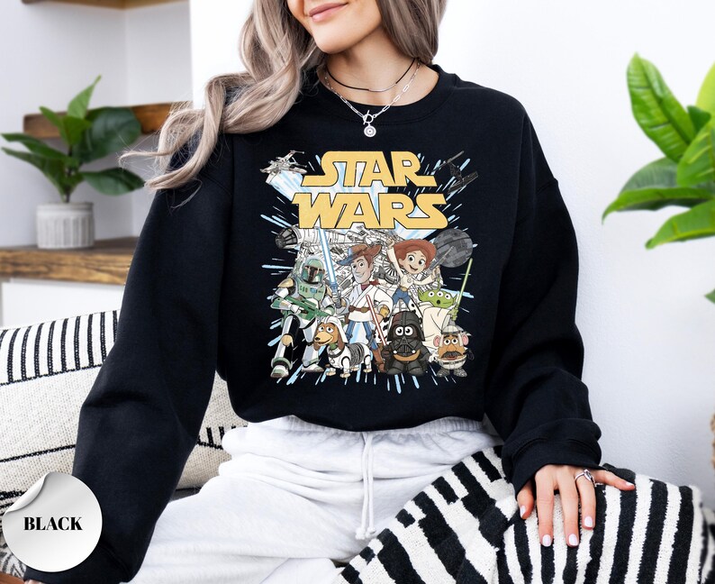 Puede incluir: Una sudadera negra de manga larga con un estampado gr&aacute;fico de personajes de Star Wars reimaginados como figuras de Toy Story. El dise&ntilde;o incluye el logotipo amarillo de Star Wars sobre ilustraciones de personajes como Woody, Jessie y Darth Vader.