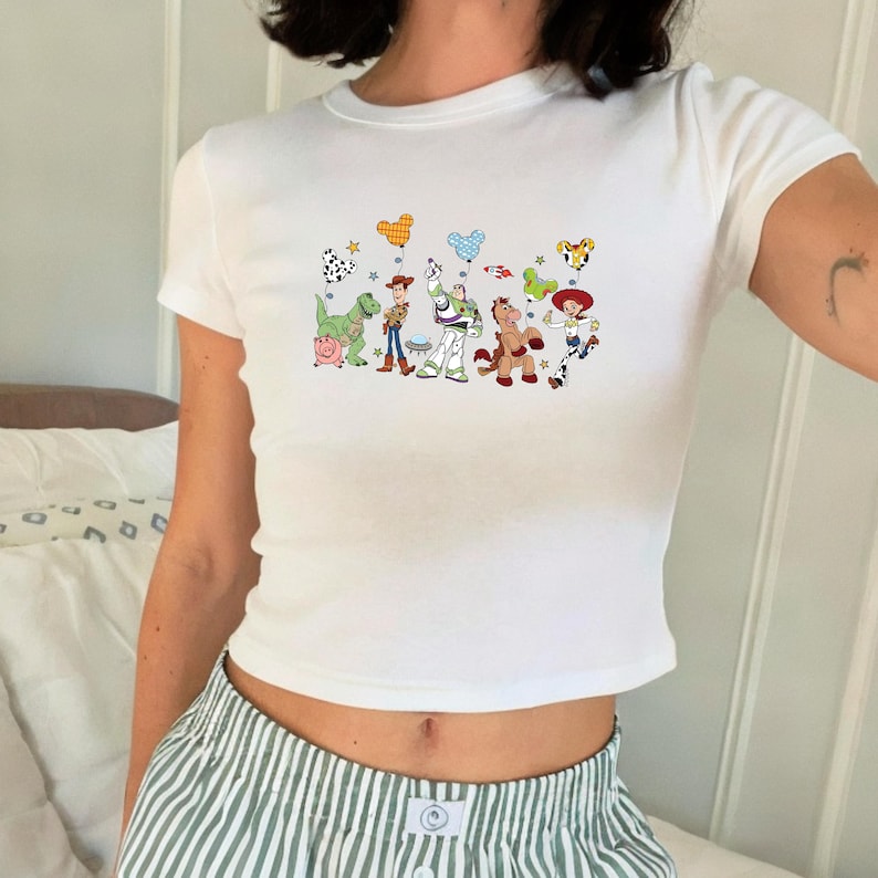 May include: A white short-sleeved t-shirt featuring a colorful graphic print of Toy Story characters including Rex, Hamm, Woody, Buzz Lightyear, Bullseye, and Jessie. The characters are holding various Mickey Mouse-shaped balloons and stars.