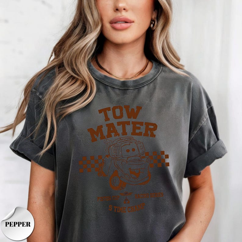 May include: A charcoal gray short-sleeve t-shirt featuring a brown graphic print of the character Tow Mater from the movie Cars. The design includes the text "TOW MATER" above the character, a checkered pattern, and additional small text below.