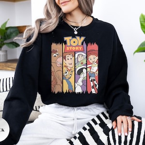 K&ouml;nnte beinhalten: Ein schwarzes Langarm-Sweatshirt mit einem Grafikdruck des Toy Story-Logos &uuml;ber vier vertikalen Feldern. Die Felder zeigen Illustrationen von Bullseye, Woody, Buzz Lightyear und Jessie in ihren charakteristischen Farben und Outfits.
