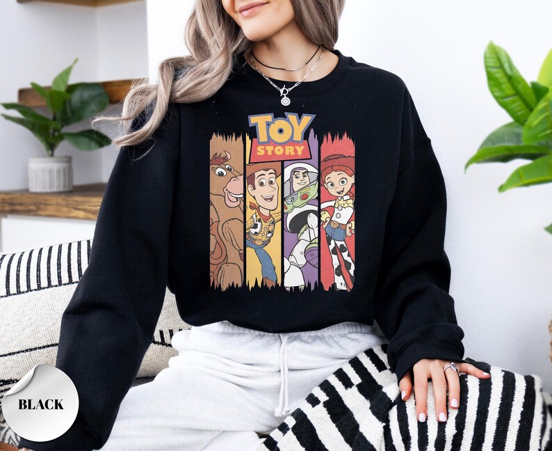 K&ouml;nnte beinhalten: Ein schwarzes Langarm-Sweatshirt mit einem Grafikdruck des Toy Story-Logos &uuml;ber vier vertikalen Feldern. Die Felder zeigen Illustrationen von Bullseye, Woody, Buzz Lightyear und Jessie in ihren charakteristischen Farben und Outfits.