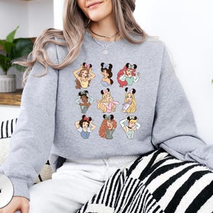 May include: A light grey sweatshirt featuring a grid of nine cartoon illustrations of princesses wearing mouse ears. Each princess has a unique hairstyle and outfit. The word "GREY" is printed on a grey circle in the lower left corner.