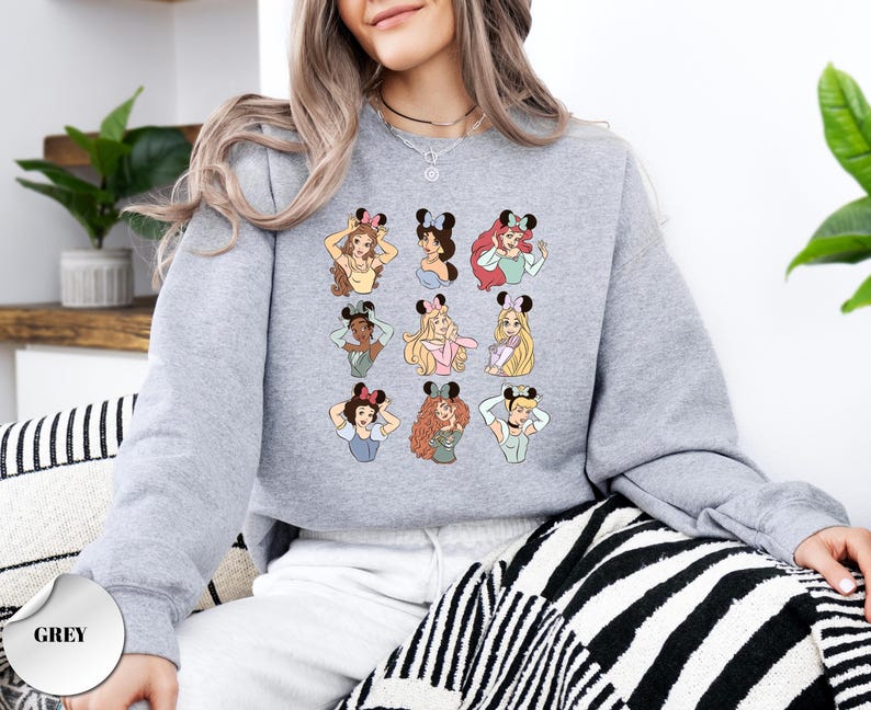 May include: A light grey sweatshirt featuring a grid of nine cartoon illustrations of princesses wearing mouse ears. Each princess has a unique hairstyle and outfit. The word "GREY" is printed on a grey circle in the lower left corner.