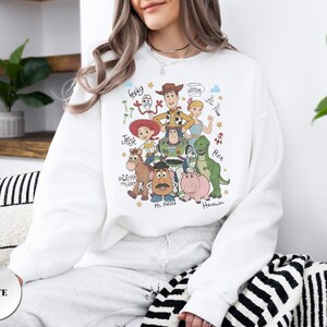 May include: A white crewneck sweatshirt featuring a colorful graphic print of Toy Story characters including Woody, Buzz Lightyear, Jessie, Bo Peep, Rex, Hamm, Mr. Potato Head, Bullseye, and Forky, with their names written in a handwritten script font.
