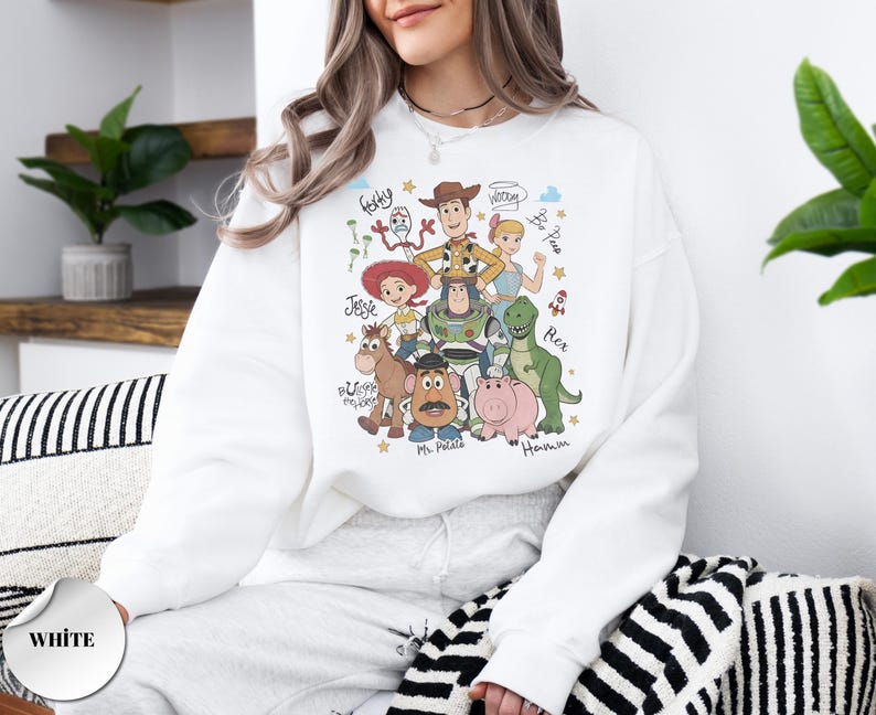 May include: A white crewneck sweatshirt featuring a colorful graphic print of Toy Story characters including Woody, Buzz Lightyear, Jessie, Bo Peep, Rex, Hamm, Mr. Potato Head, Bullseye, and Forky, with their names written in a handwritten script font.