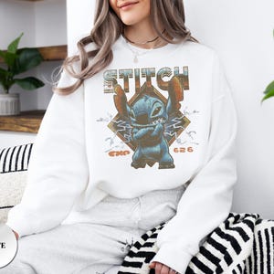 May include: A white crewneck sweatshirt featuring a graphic print of the blue character Stitch with arms crossed. The design includes the text STITCH in a distressed font, a diamond-shaped background, lightning bolts, and the text EXP 626.